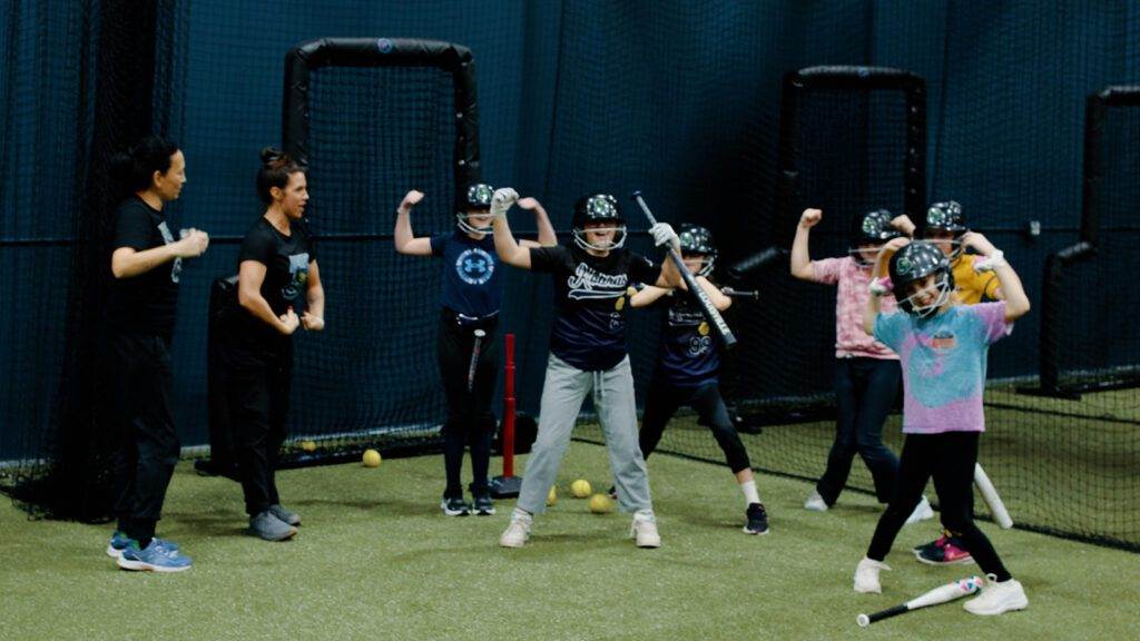 A softball coach and her team playfully flexing while in the batting cage